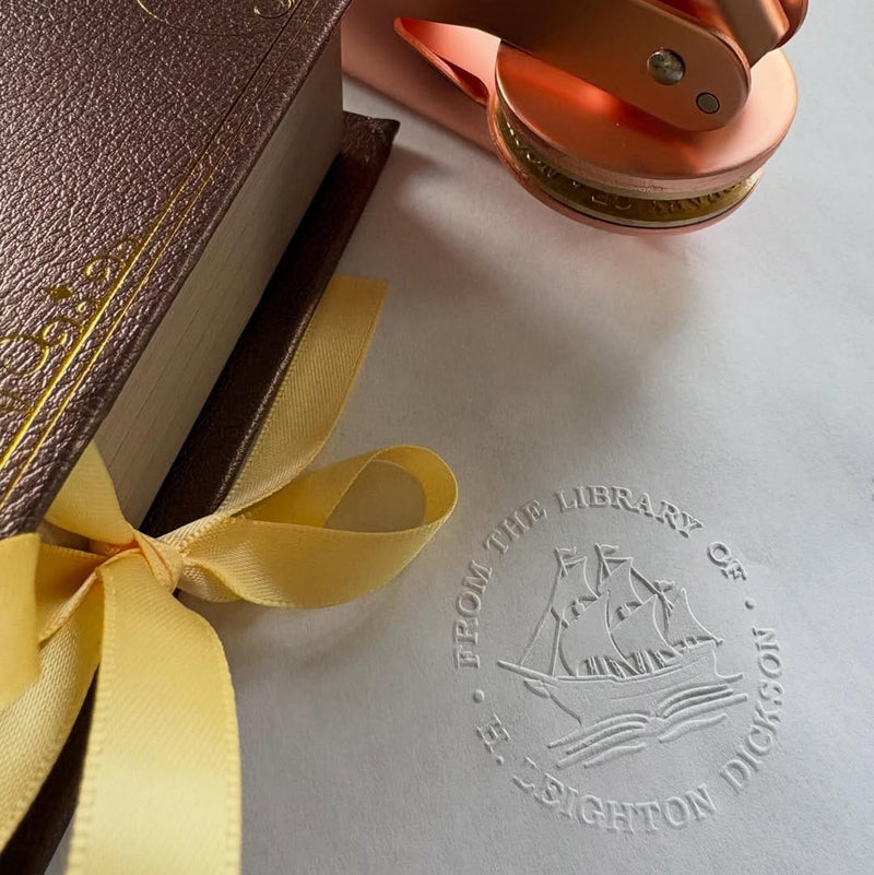 Brown leather-bound book with a yellow ribbon and orange metal object on a gray surface with a ship logo.
